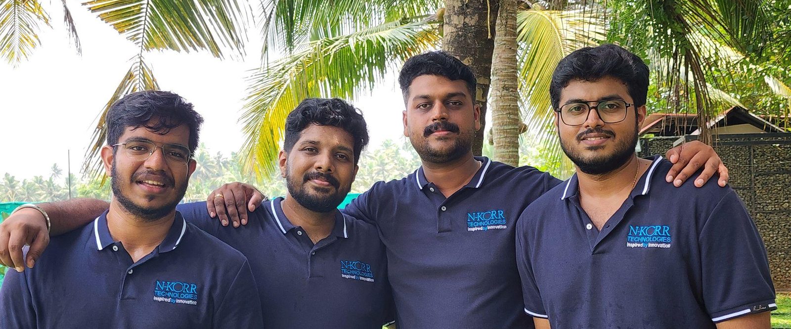 Reference image from the dataset: a group photograph of four colleagues in matching NKORR polo shirts standing outdoors in front of palm trees.